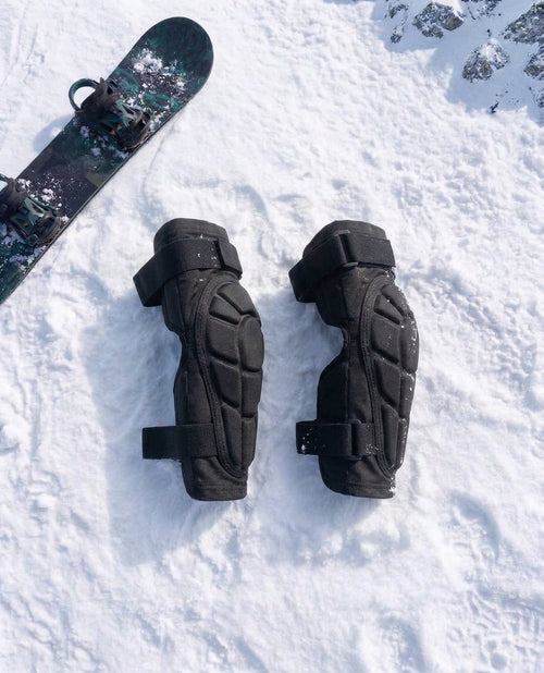 Pactline snow elbow pads laid flat on snow beside a snowboard